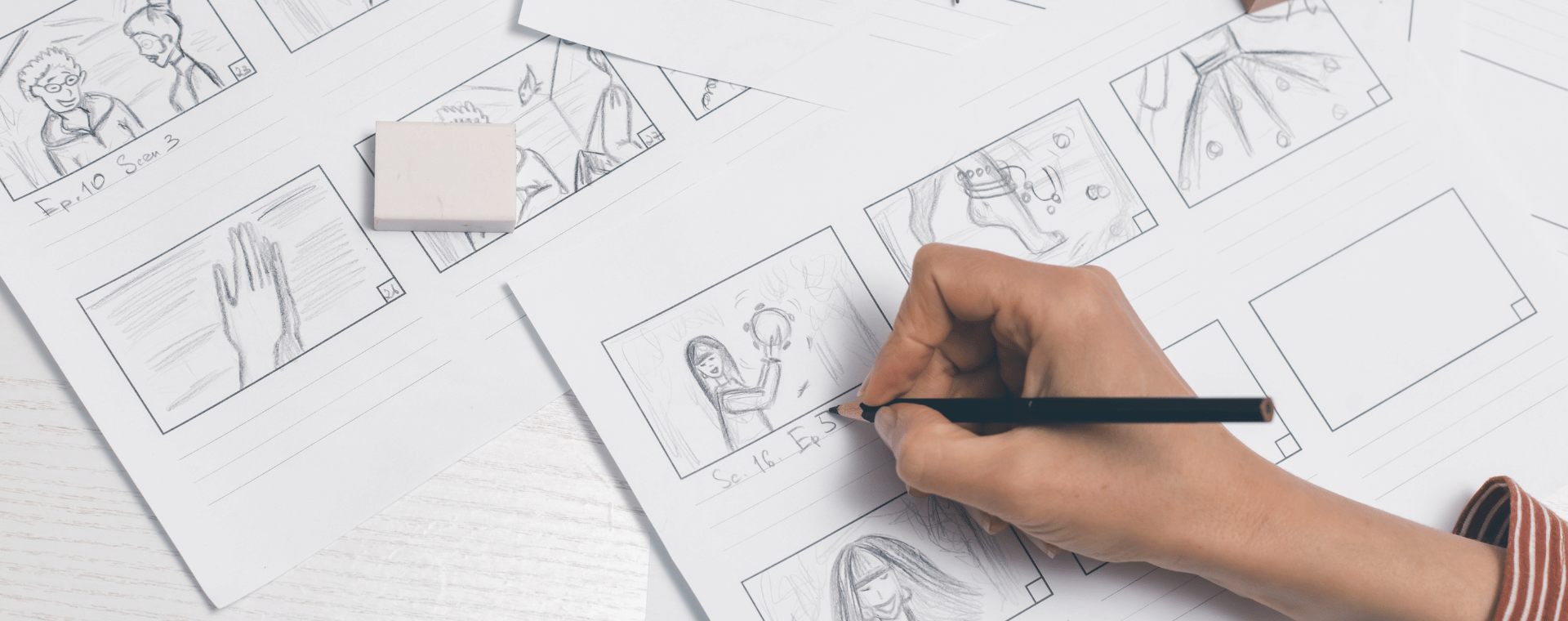 A person's hand is drawing storyboards for an animation on paper with a pencil. Several sheets with rough sketches and scenes, all part of the promotional video, are scattered on the desk. An eraser is placed among the sheets.