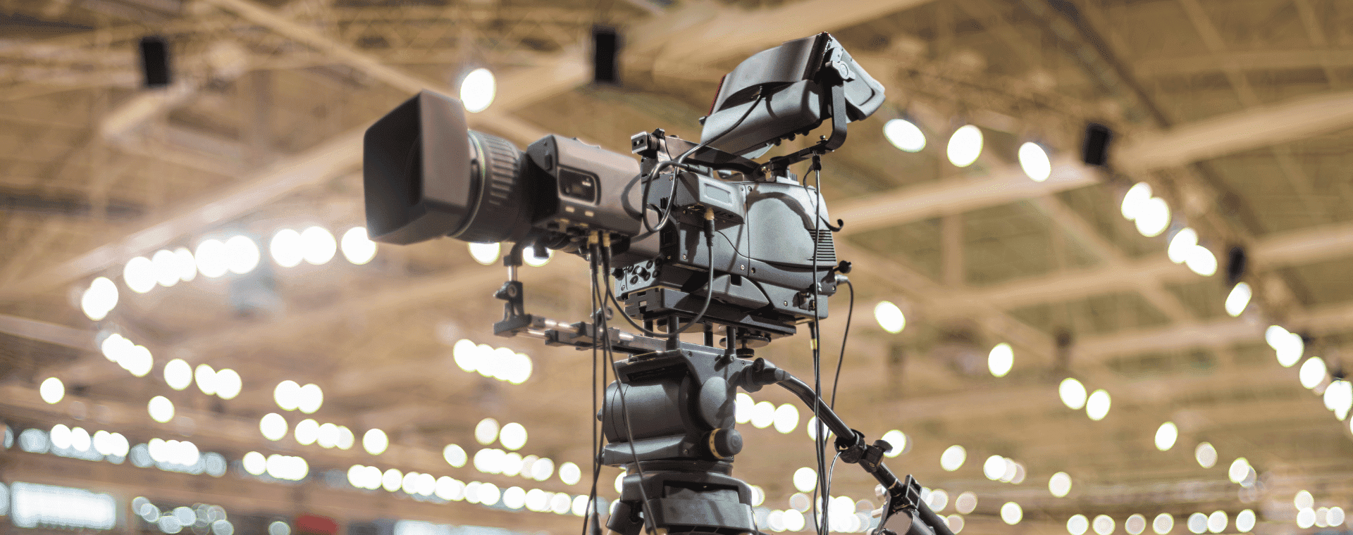 A professional 4K video camera is set up on a tripod, focusing on an indoor scene with blurred background lights, suggesting a large venue. The camera, one of the best yet cheapest options for high-quality filming, is equipped with various attachments and appears ready for broadcasting.