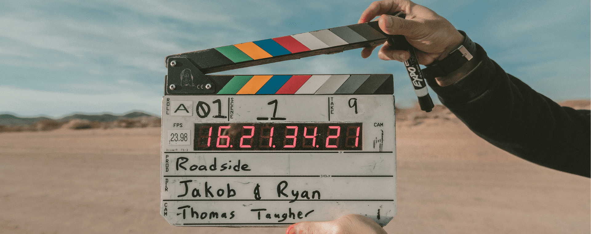 A person holds a clapperboard in front of an outdoor desert scene, capturing footage with one of the cheapest 4k cameras. The clapperboard displays the text: "Roadside, Jakob & Ryan, Thomas Taugher" and various numerical data, including "16:21:34:21". The board's top edge has colorful stripes.