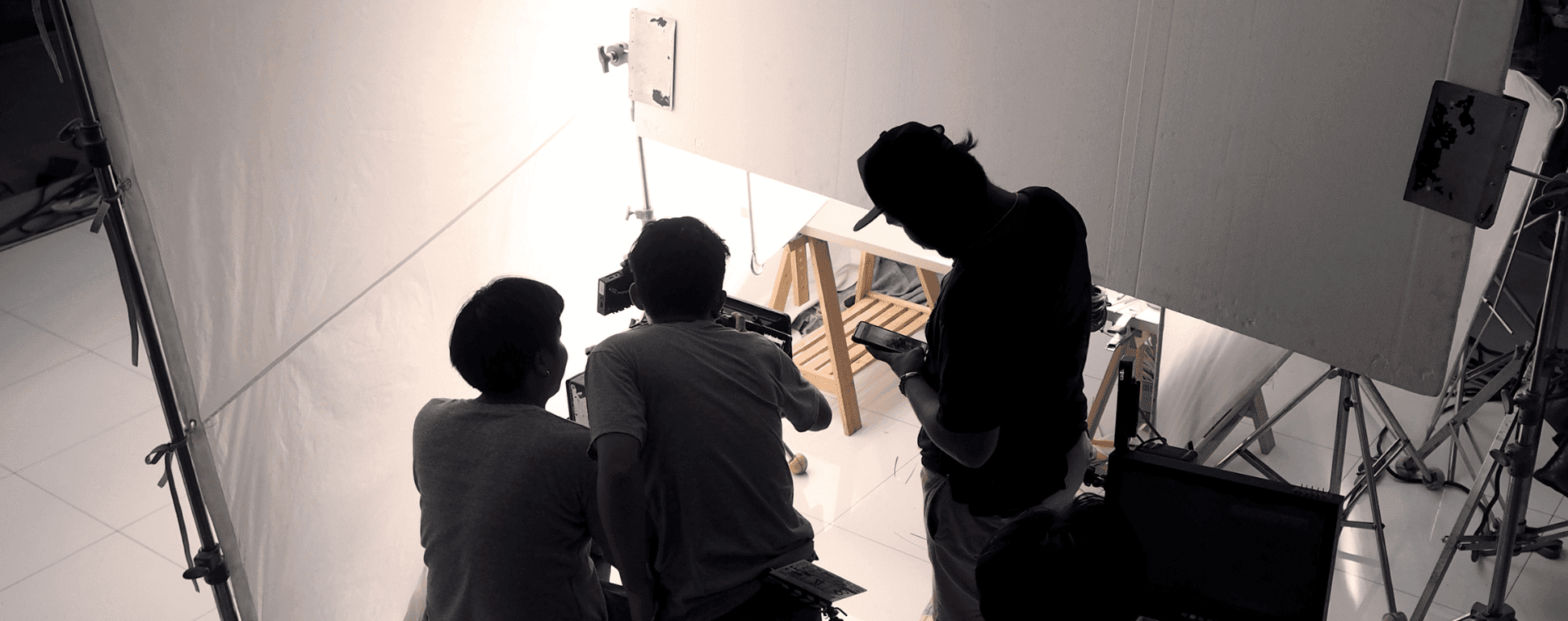 Three people are engaged in a photoshoot or filming session, using professional cameras to capture footage in stunning 4K. They are looking at a camera setup aimed at a small wooden chair on a white background, surrounded by professional lighting equipment and reflectors. The scene suggests a controlled studio environment.