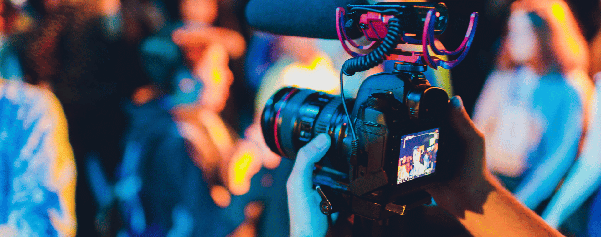 Close-up shot of a person holding a professional video camera with a mounted microphone, recording a group of people in a brightly lit, blurred background. The scene captures the magic of event filming with vibrant lighting and dynamic atmosphere.