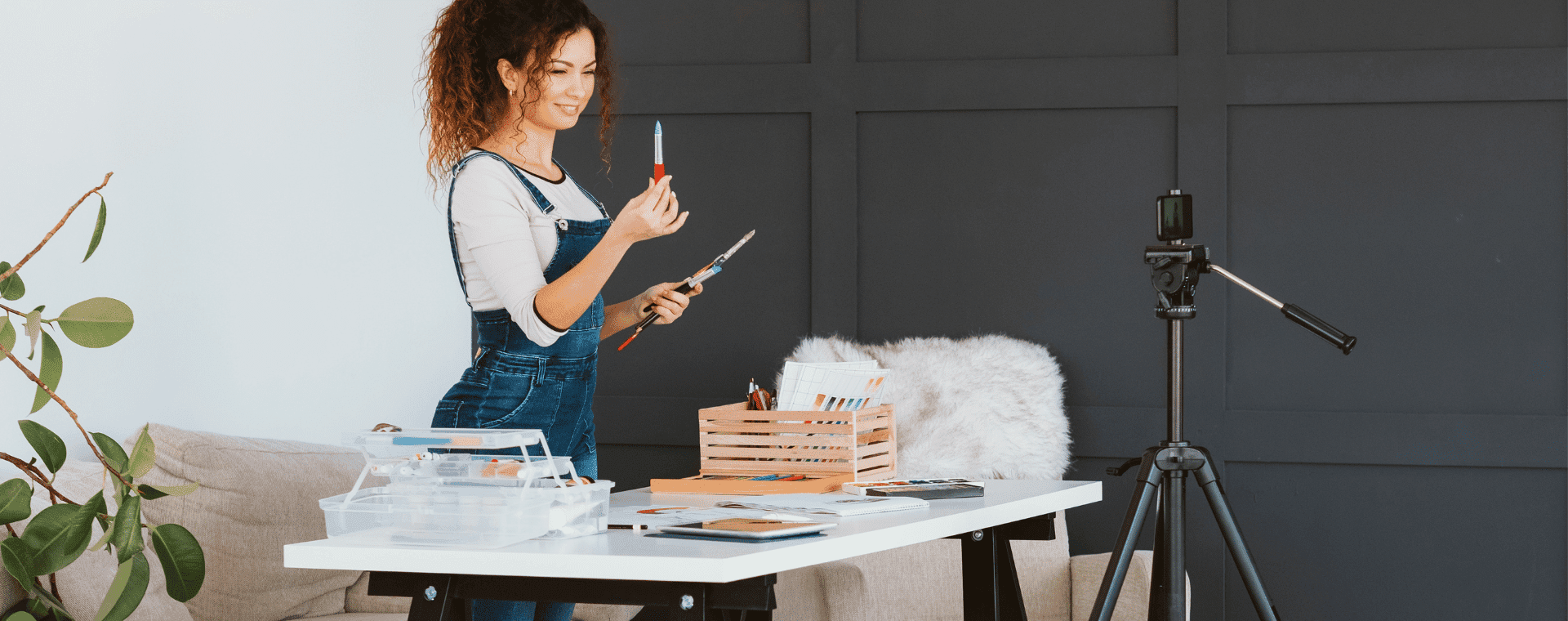 A woman wearing overalls stands at a desk, holding art supplies. The desk is covered with various art tools and materials. A professional 4k camera on a tripod is positioned in front of the desk, suggesting she might be recording a video. There is a plant and a couch in the background.