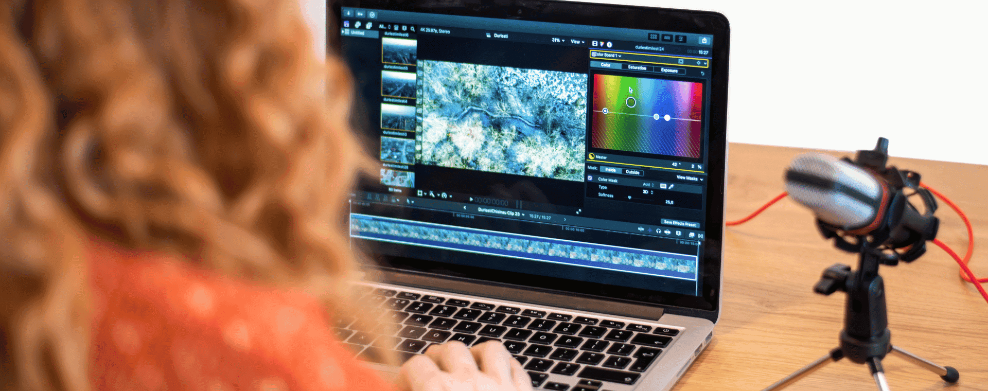A person with curly hair, seen from behind, works on video editing on a laptop. The screen displays a video timeline and color correction tools. A microphone on a stand is visible to the right of the laptop, and in the background, there are professional cameras for multimedia production.