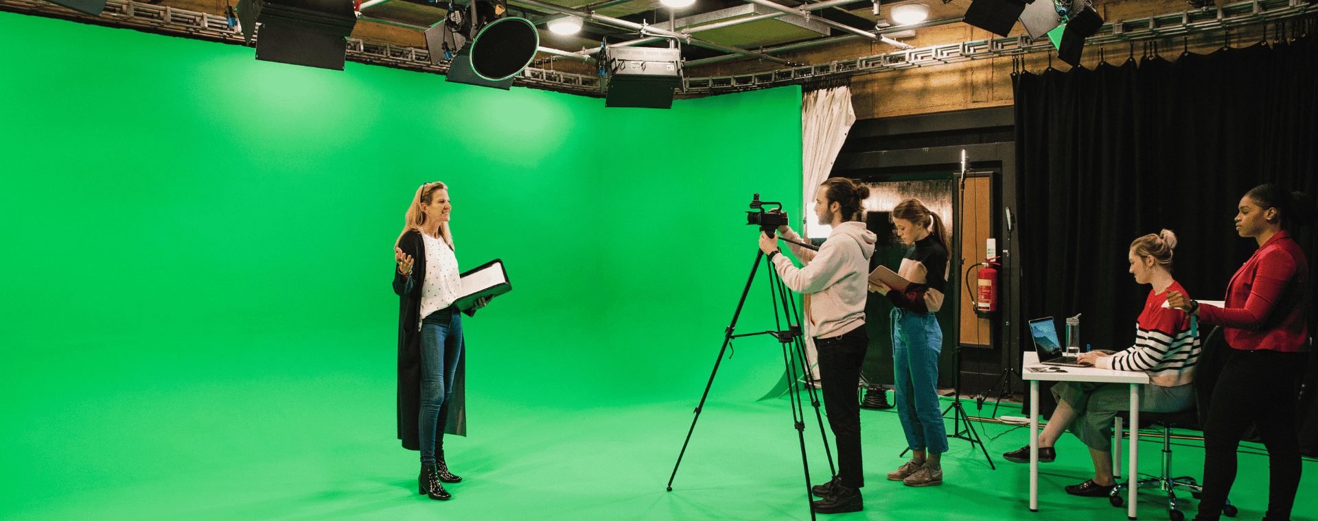 A woman stands in front of a green screen, holding a tablet and speaking to create an effective video. Four people assist with camera and audio equipment while two others work at a nearby table with a laptop. The setting is a professional studio designed for optimal SEO content creation.