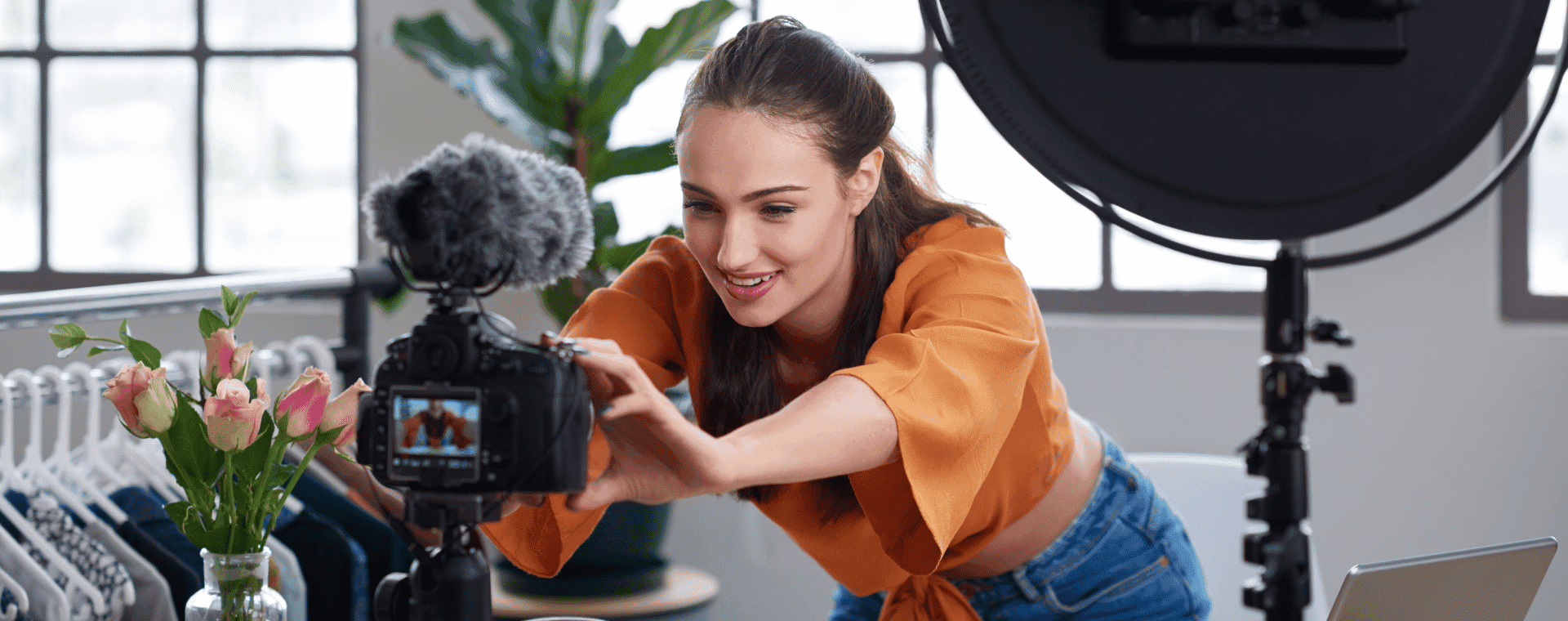 A woman wearing an orange blouse adjusts a camera on a tripod surrounded by filming equipment, including a ring light. In the background, clothing items hang on a rack and a vase of flowers sits on a nearby table, all set for creating content that ensures her video gets seen.