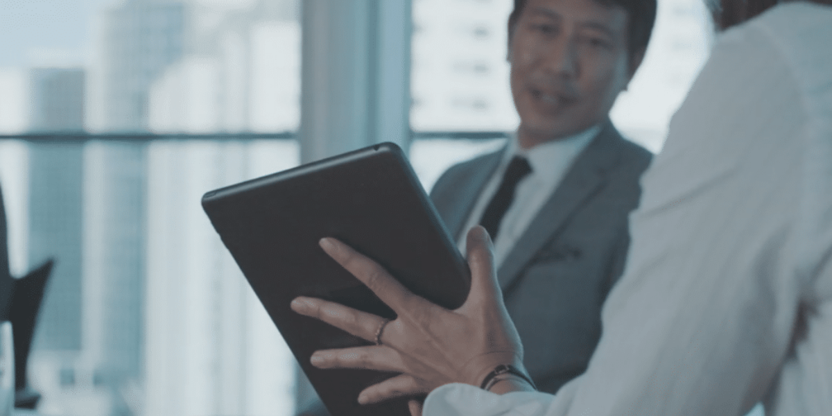A close-up of a person's hand holding a tablet, showing the back of the device. In the background, a man in a suit is slightly blurred and facing the person with the tablet. The office setting has large windows with a cityscape view; they are likely discussing features of the Cinema Forms APP.