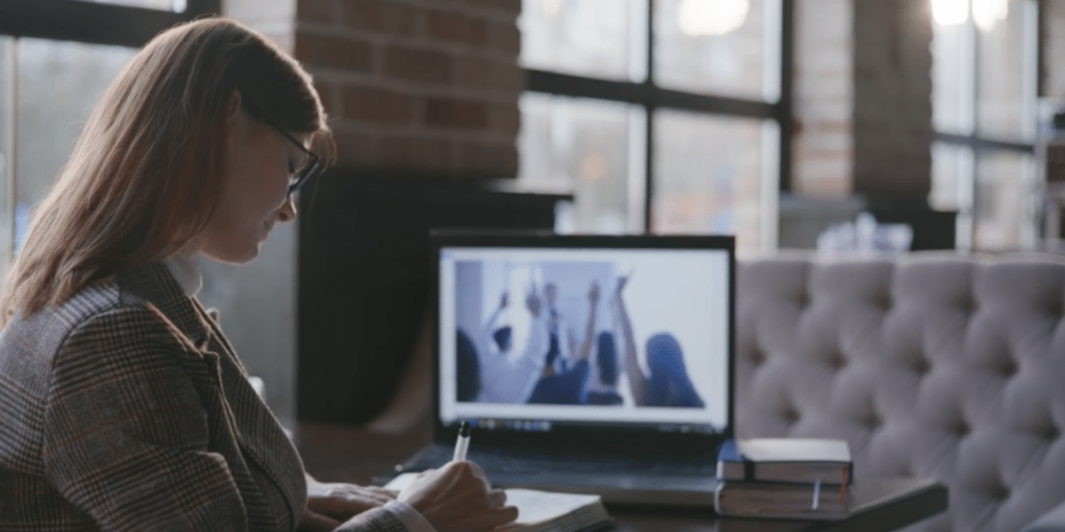 A person sits at a table with a notebook, writing. They face a laptop displaying a virtual meeting where participants appear to be raising their hands. The room's large windows and brick walls suggest it’s a modern workspace typical for an SEO-focused professional.