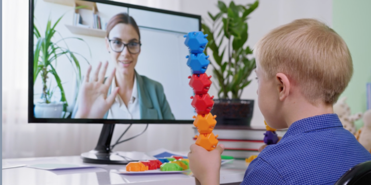 A young boy sits at a desk holding a stack of colorful interlocking toys while video chatting with a woman on a computer screen. The woman, wearing glasses and a suit jacket, is smiling and waving. The background shows a bright room with plants and shelves, creating an engaging learning environment.