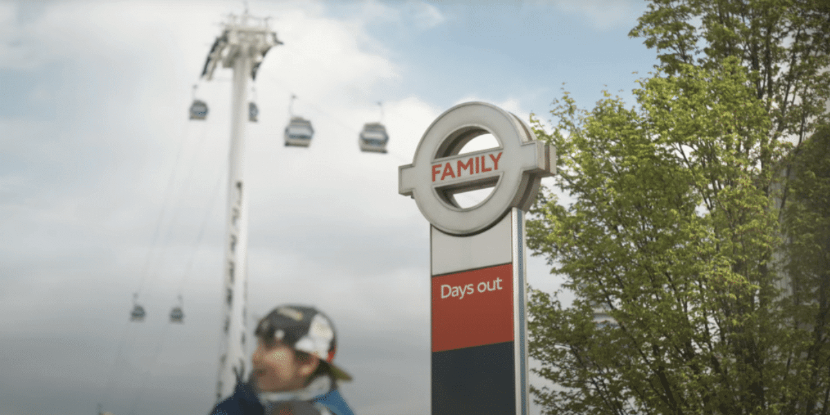 A signpost with the words "FAMILY" and "Days out" stands near some trees, hinting at a perfect destination for a creative day. In the background, cable cars glide on a line while a person in a hat and sunglasses walks by. The partly cloudy sky adds to the scenic charm, perfect for video advertising.