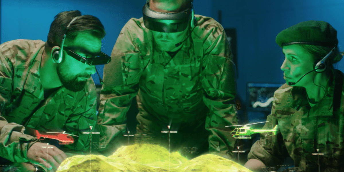 Three military personnel in camouflage uniforms are observing and interacting with an animated compositing of a holographic battlefield map. They are wearing headsets and augmented reality visors. Two helicopters, one red and one white, are projected on the map above the terrain.