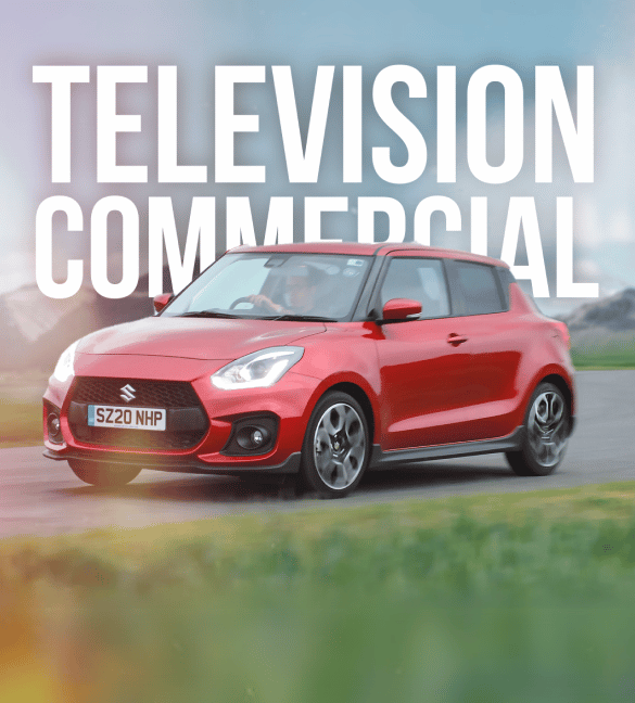 A red Suzuki car with a license plate "SZ20 NHP" is shown driving on a road against a mountainous background. Bold white text above the car reads "TELEVISION COMMERCIAL." The road and foreground are slightly blurred to imply motion, capturing the essence of our video production services in action.