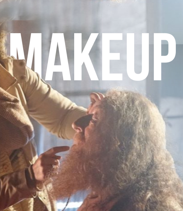 A person with long hair and a beard is sitting while another, partially visible, applies makeup to their face. Large white text saying "MAKEUP" is prominently displayed in the upper part of the image, suggesting a professional video production service at work.