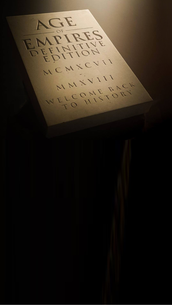 A white book titled "Age of Empires Definitive Edition" is displayed against a dark background. The cover features Roman numerals MCMXCVII and MMXVIII, and the text "Welcome Back to History" at the bottom. The dim lighting, reminiscent of professional video production services, adds a dramatic effect.
