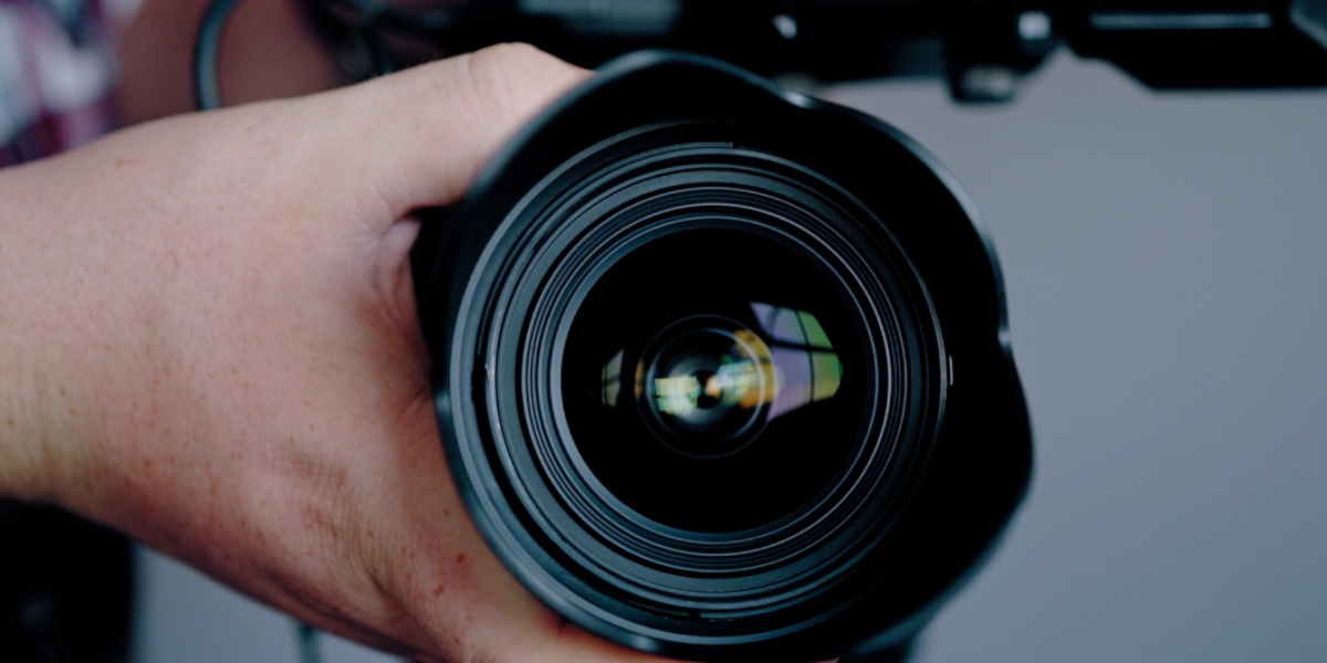 Close-up of a person holding a DSLR camera pointed directly at the viewer, perfect for video content creation. The lens is central in the frame, with a visible reflection on the glass, showing intricate details of the hand and lens structure. The background is blurred.