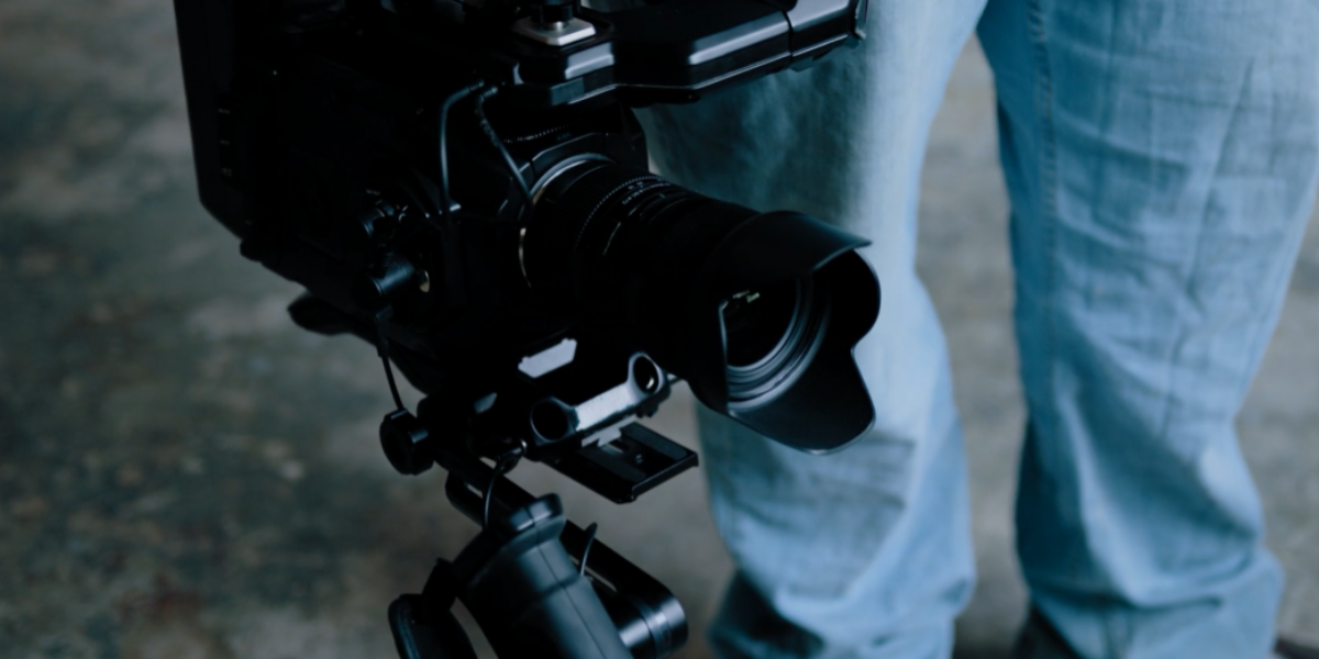 Close-up of a person using a professional camera mounted on a tripod, capturing brand video production: maximizing your brand's potential. The individual's lower torso and jeans are visible. The camera has a large lens and additional accessories attached, set indoors against neutral flooring.