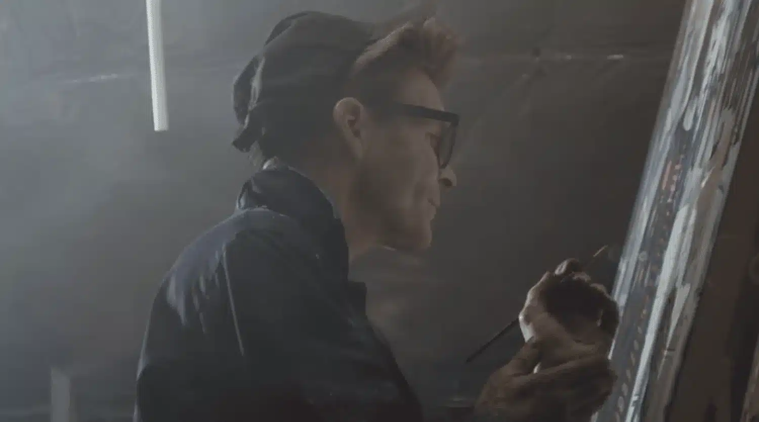 A Person, Wearing Glasses And A Dark Cap, Is Side-Facing And Painting On A Canvas In What Appears To Be Footage For A Promotional Video. The Background Is Somewhat Hazy, And They Hold A Paintbrush, Seemingly Focused On Their Artwork. The Dim Lighting Adds An Atmospheric Ambiance To This Artistic Case Study. A person, wearing glasses and a dark cap, is side-facing and painting on a canvas in what appears to be footage for a promotional video. The background is somewhat hazy, and they hold a paintbrush, seemingly focused on their artwork. The dim lighting adds an atmospheric ambiance to this artistic case study.