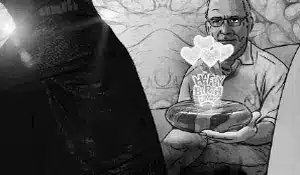 A grayscale image shows an older man in glasses sitting and holding a bowl with flowers and a stick that reads "Happy Birthday" sticking out of it. In the foreground, a person dressed in dark clothing partially obstructs the view, reminiscent of vintage video advertising.