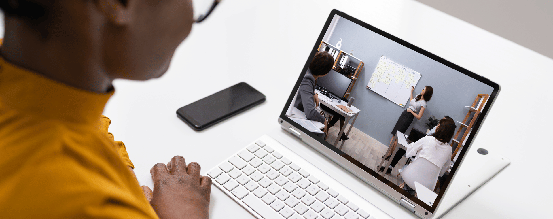A person sits at a desk, looking at a laptop screen showing a training video production. The video displays a meeting setting with three people; one is standing and pointing at a whiteboard, while two others are seated and watching attentively.