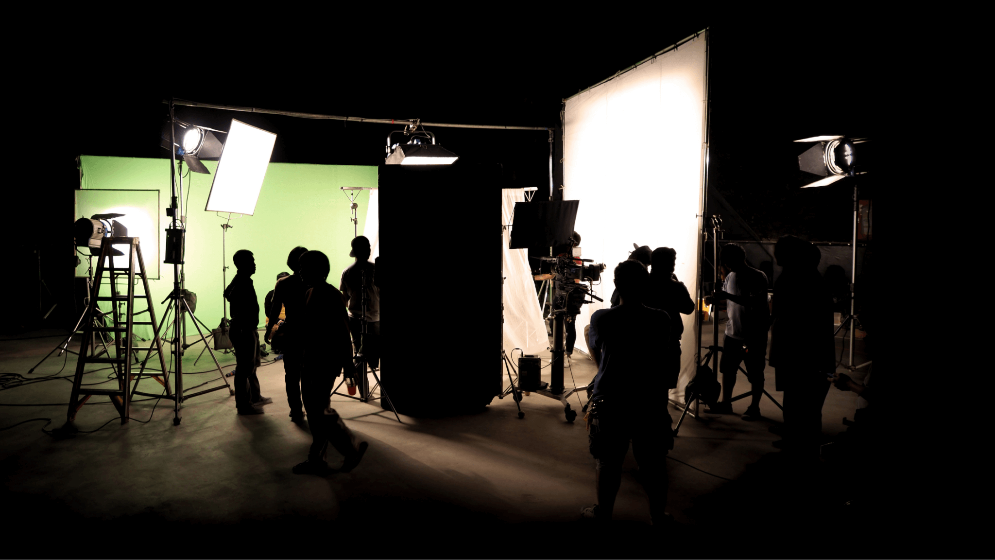 A busy film set with several crew members working around various lighting and filming equipment. A green screen and several bright lights illuminate the scene. Silhouetted figures provide an energetic vibe as they prepare for shooting, reminiscent of top-notch video production companies near you.
