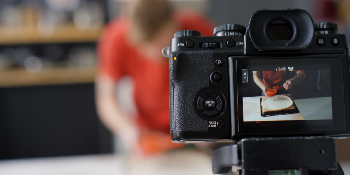 A camera is positioned on a tripod, capturing a person in a red shirt preparing food at a counter. The camera's screen displays a close-up view of the person working with a baking tray, perfect for video marketing content. The background is blurred.