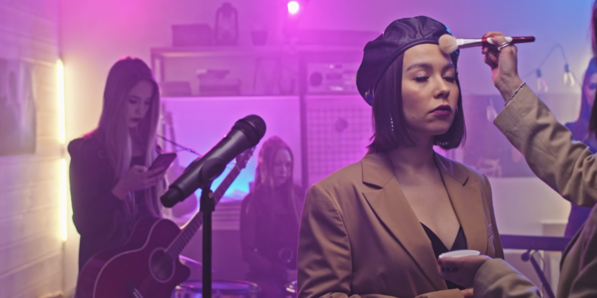 A woman in a beige blazer and black beret gets makeup done while standing in front of a microphone. Another woman in the background is tuning a guitar and checking her phone, capturing the hustle of music video production. The scene is lit with a pink and purple hue, with music equipment visible around them.