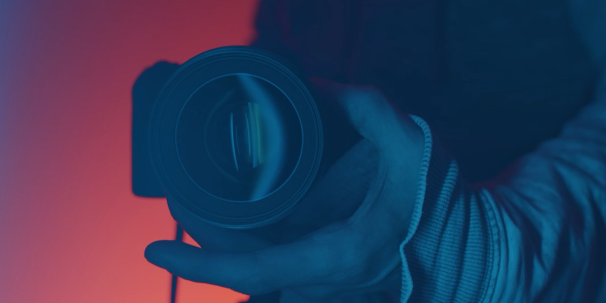 A close-up of a person holding a camera lens with one hand. The background features a gradient of red and blue colors, creating a dramatic lighting effect, reminiscent of video post-production services. The person's arm is covered with striped long sleeves.