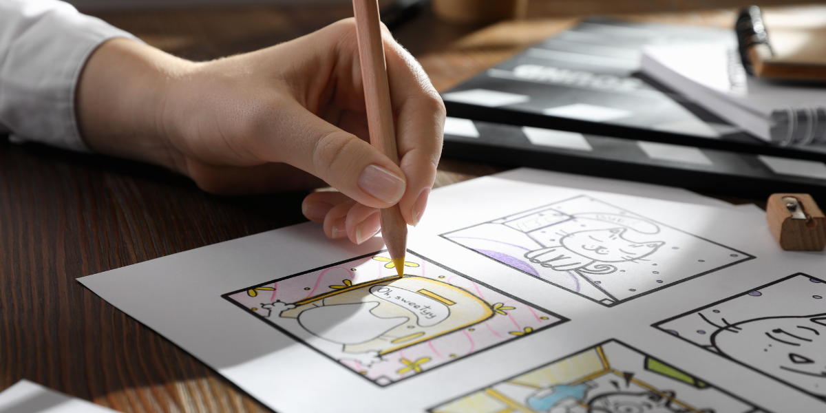 A person's hand is shown drawing or coloring a comic strip with color pencils on a white sheet of paper. The comic strip features a character wearing a hat and various scenes. Other drawing and writing tools, including a sharpener, are scattered on the wooden desk, as if preparing for a creative video.
