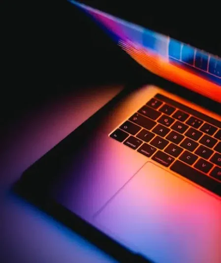 A close-up image of a laptop with its screen partially closed, casting vibrant, colorful light reflections on the keyboard. The hues range from deep blues and purples to bright oranges and yellows, creating a striking and modern visual effect, perfect for video production resources.