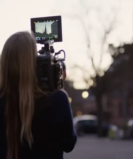 A person with long hair stands outdoors, holding and filming with a professional video camera. The camera's screen displays an image of a building with distinct architecture. The background is blurred with soft lighting and silhouettes of trees and buildings, a perfect scene for tapping into various video production resources.