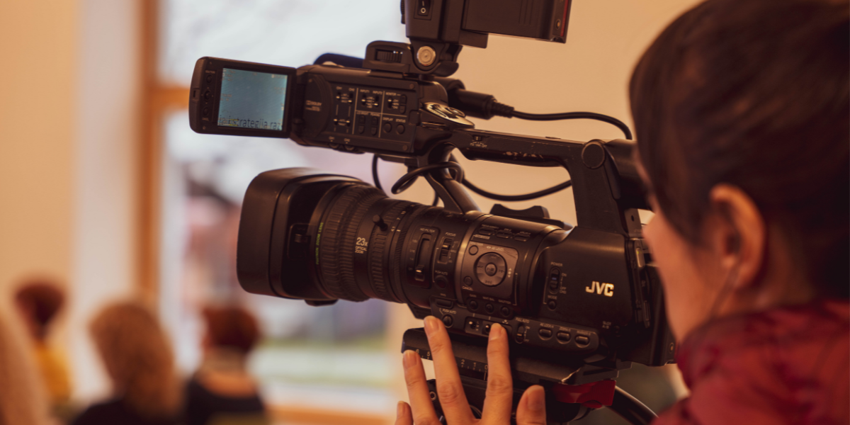 A person operates a professional JVC video camera, pointing it towards a group of blurred individuals in the background. The setting, bathed in natural light streaming through a window, suggests an engaging indoor scene for a promotional video.