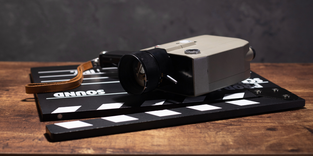 A vintage film camera with a black lens and a brown strap rests on a black and white movie clapperboard labeled "SOUND." The scene evokes the artistry of screenwriting, set on a wooden surface against a dark, textured background.