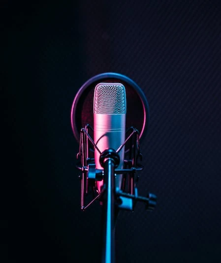 A close-up image of a professional condenser microphone with a pop filter in a dimly lit recording studio. The microphone is illuminated by purple and blue lighting, creating a vibrant, atmospheric effect. Ideal for video production resources, the background is dark and textured.