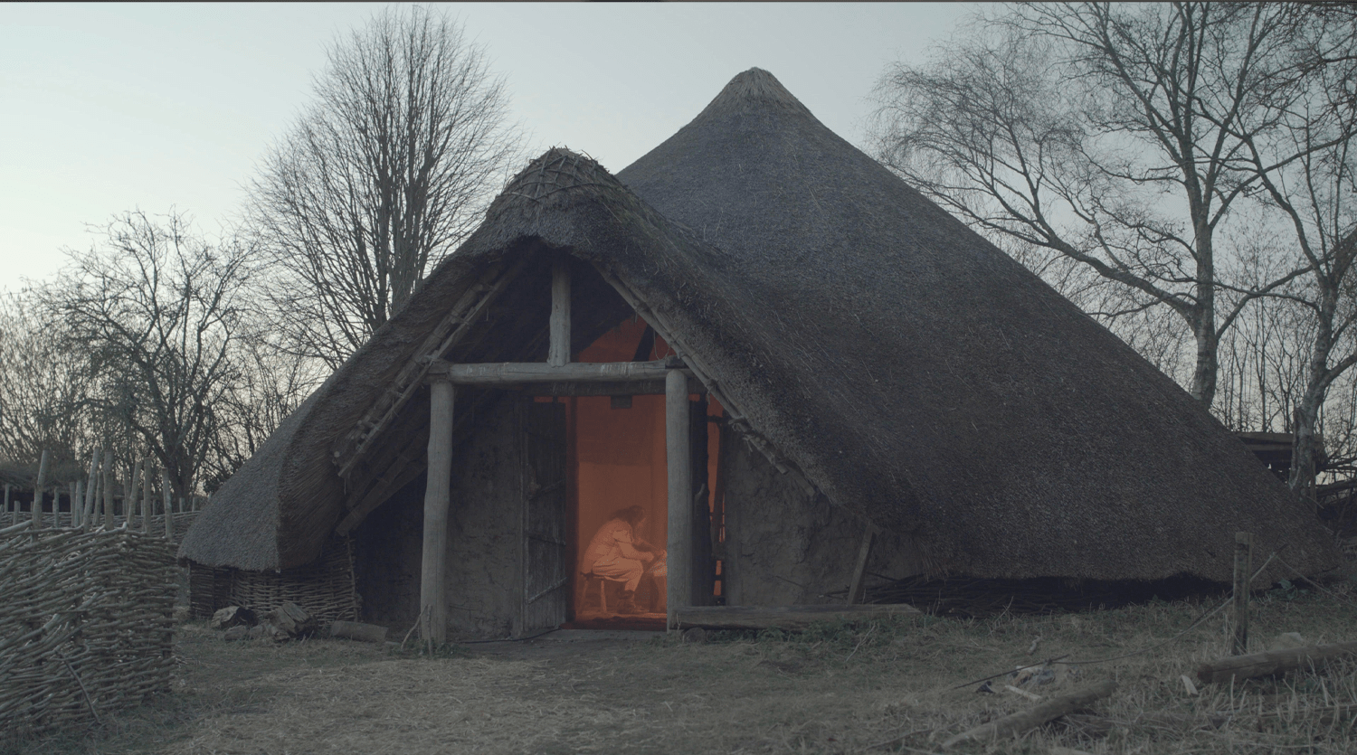 An ancient thatched-roof structure stands surrounded by bare trees and a wicker fence. Inside, a warmly lit area reveals a person sitting, partially illuminated through an open doorway. The serene, historic setting provides an ideal backdrop for video production services under the dim sky.