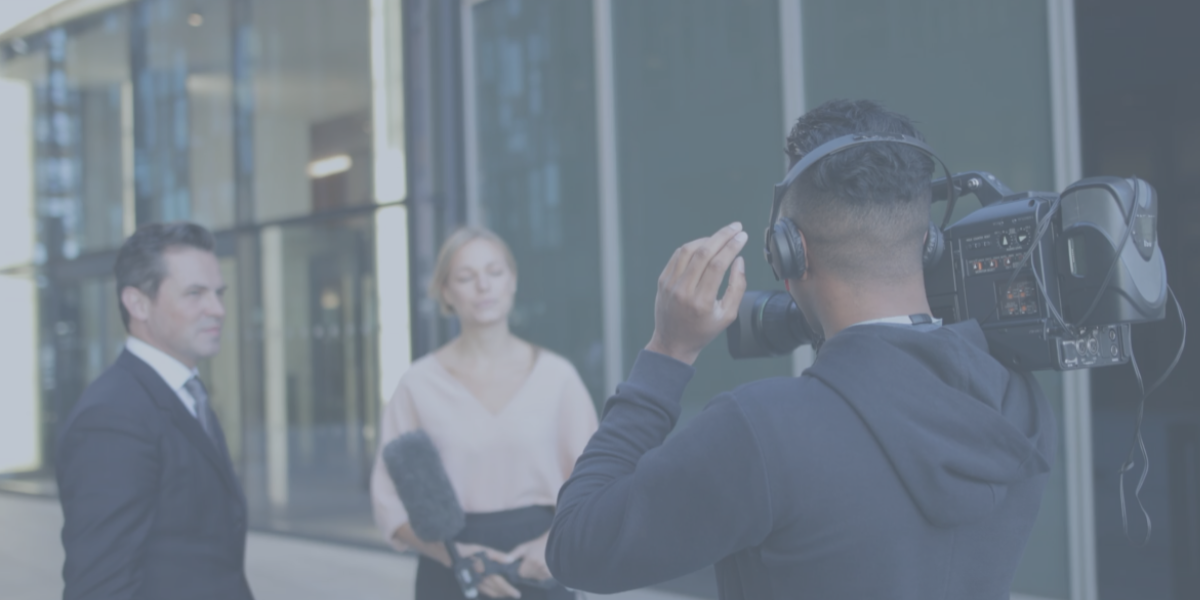 A cameraman wearing headphones films a man in a suit and a woman holding a microphone. They are standing outside a modern glass building, appearing to be in the middle of filming corporate videos for an interview.