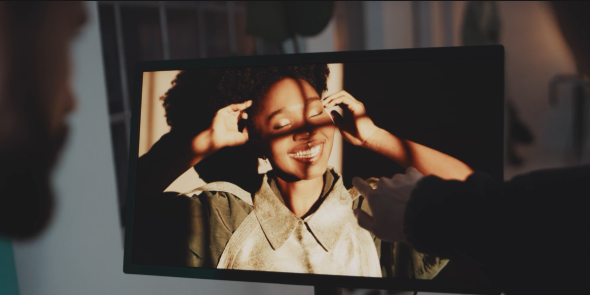 A person points at a computer screen displaying a photograph of a smiling person with short curly hair. The subject in the photo, perfect for marketing campaigns, stands in sunlight, using their hands to shield their face from the light. The monitor's frame is partially visible.