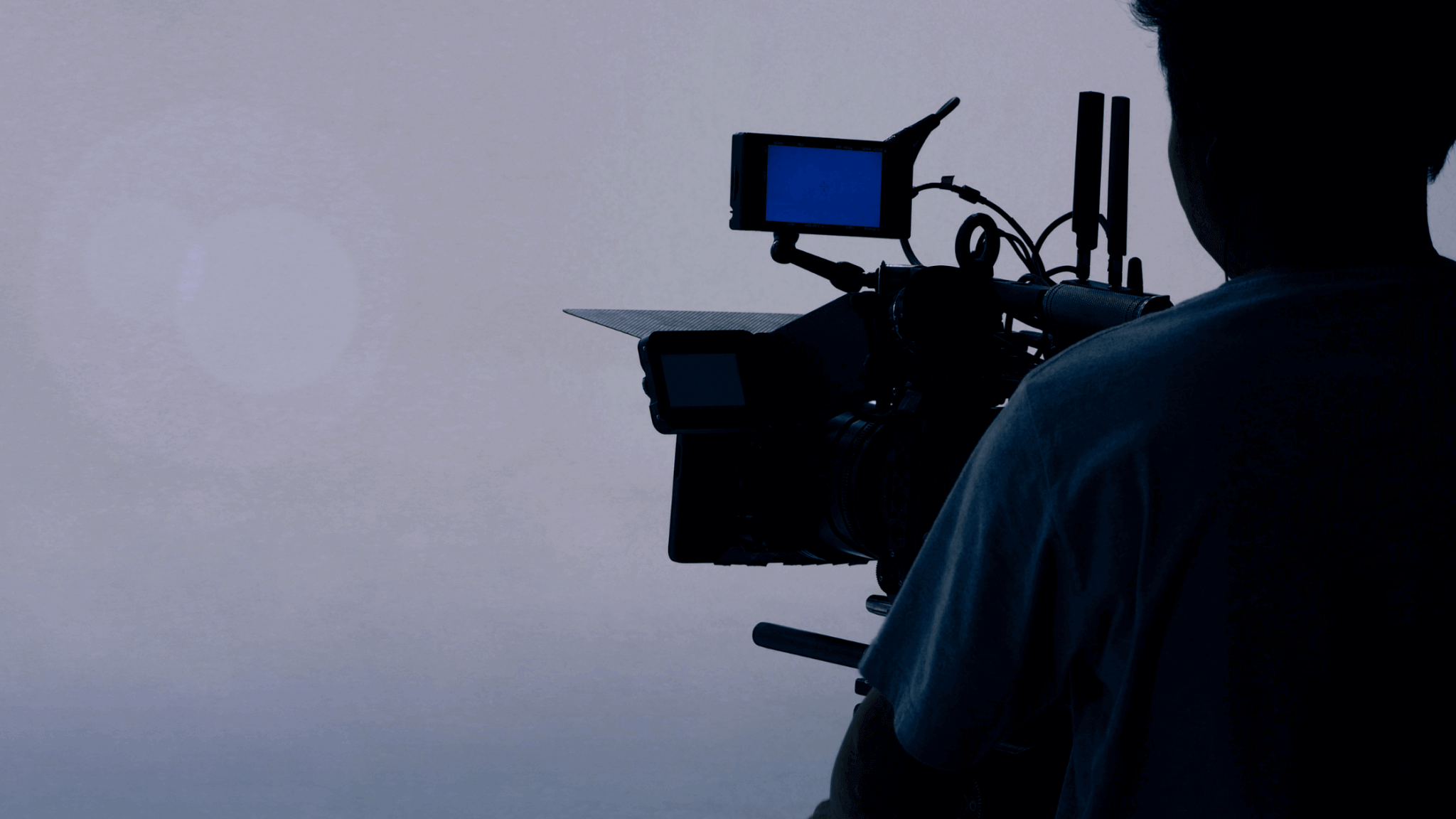 A silhouette of a person operating a professional video camera with a small monitor attached, capturing the perfect shot for a promotional video. The background is a gradient of light to dark blue, creating a serene, minimalist atmosphere.