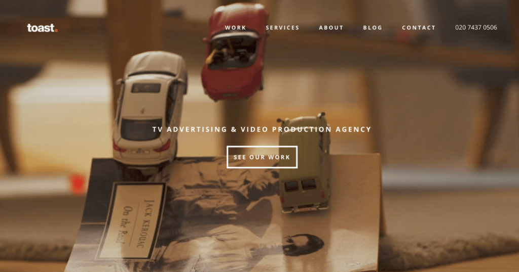 A Desktop Scene With Miniature Cars On A Stack Of Vintage Items, Including A Book Titled &Quot;On The Road&Quot; By Jack Kerouac. The Website Menu At The Top Includes Options: Work, Services, About, Blog, And Contact. Discover Why We'Re Among The Top 10 Video Production Companies With Our &Quot;See Our Work&Quot; Button Prominently Displayed. A desktop scene with miniature cars on a stack of vintage items, including a book titled "On the Road" by Jack Kerouac. The website menu at the top includes options: Work, Services, About, Blog, and Contact. Discover why we're among the Top 10 Video Production Companies with our "See Our Work" button prominently displayed.