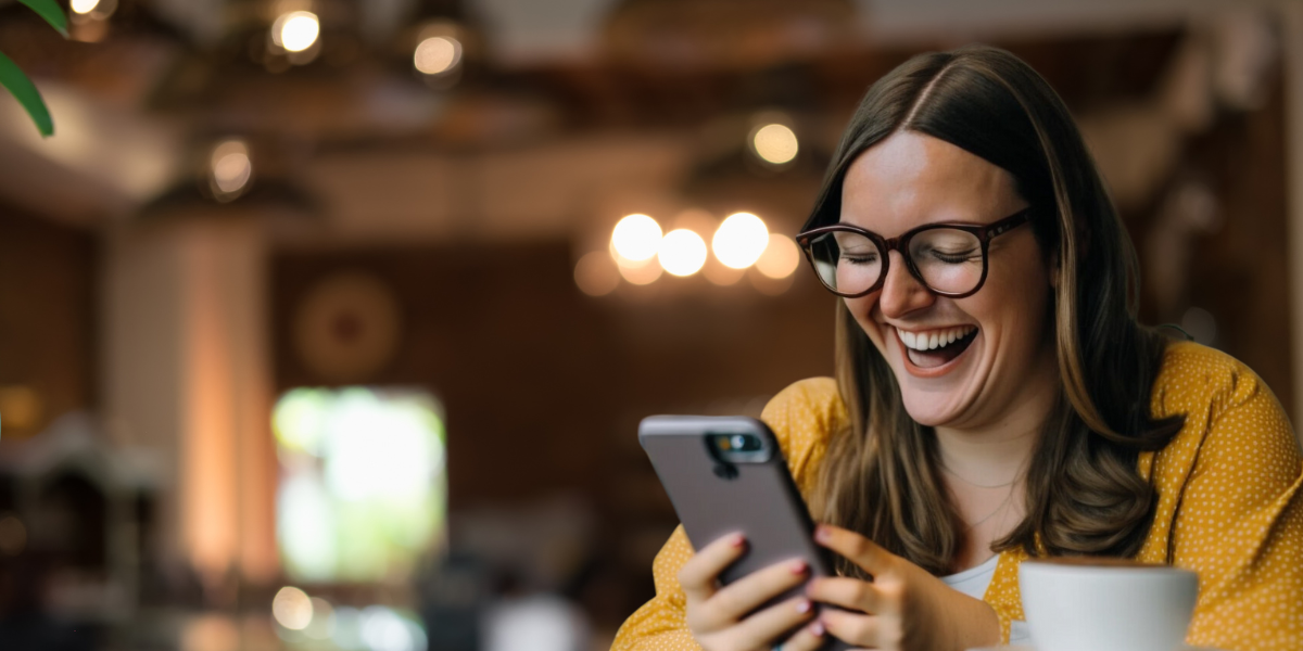A woman with glasses and long hair, wearing a yellow top, is sitting indoors and smiling while looking at her smartphone. She is holding the phone with both hands and appears to be very happy or amused, possibly watching video content. A cup sits in front of her on the table.