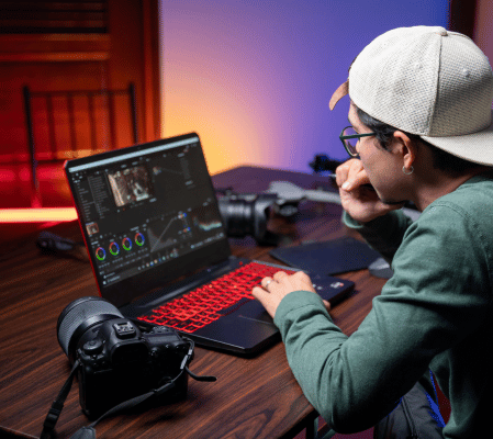 A Person Wearing A Light-Colored Baseball Cap And Glasses Is Intently Working On A Photo Or Video Editing Program On A Laptop With A Red Backlit Keyboard. Multiple Cameras, Popular Tools Among Video Editors, And A Smartphone Are Also On The Wooden Table. The Background Is Softly Lit. A person wearing a light-colored baseball cap and glasses is intently working on a photo or video editing program on a laptop with a red backlit keyboard. Multiple cameras, popular tools among video editors, and a smartphone are also on the wooden table. The background is softly lit.
