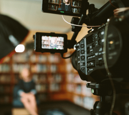 A Professional Video Camera Is Focused On A Person Sitting With Blurred Bookshelves In The Background. The Camera’s Viewfinder Shows The Person Being Recorded, Highlighting The Live-Action Video Style And Indicating An Indoor Setting, Possibly A Library Or Study. A professional video camera is focused on a person sitting with blurred bookshelves in the background. The camera’s viewfinder shows the person being recorded, highlighting the live-action video style and indicating an indoor setting, possibly a library or study.