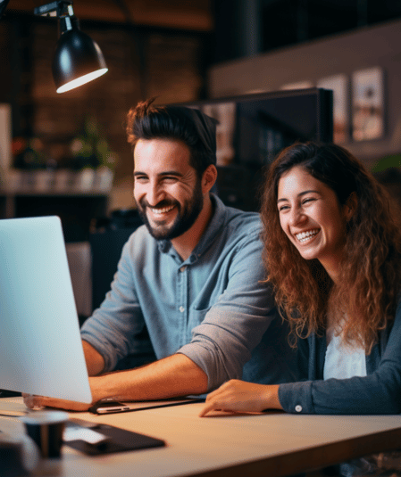 Two young adults, a man and a woman, are sitting at a desk, smiling and looking at a computer screen. The man has a beard and is wearing a blue shirt, while the woman has curly hair and is wearing a gray sweater. The setting appears to be an office environment where they are exploring websites to upload your video content.