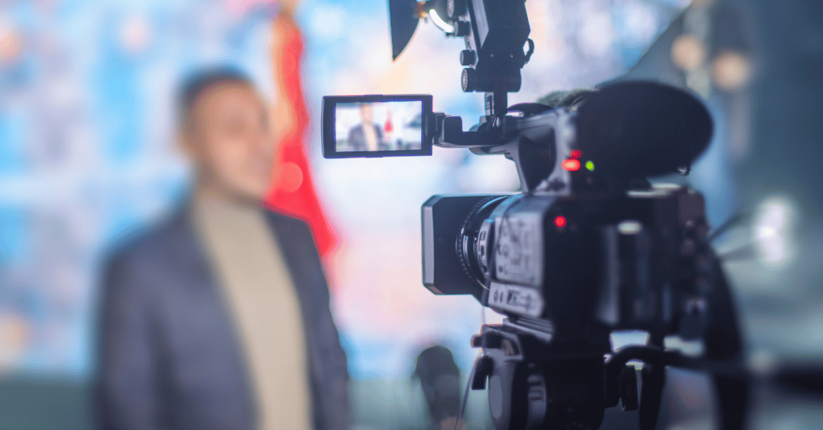 A close-up of a video camera recording a person standing in front of a blurred, colorful background. The camera's screen displays a clear image of the person, creating an essential shot for video edit purposes. The setting appears to be indoors, possibly at an event or studio. The person is partially out of focus.