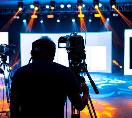 A Silhouetted Camera Operator Films In A Studio With Vibrant Lighting. The Background Features Large Led Screens Displaying White Content, And The Stage Is Illuminated With Blue And Orange Lights, Creating An Energetic Atmosphere That Blurs The Lines Between Animation Vs Live-Action Video Styles. A silhouetted camera operator films in a studio with vibrant lighting. The background features large LED screens displaying white content, and the stage is illuminated with blue and orange lights, creating an energetic atmosphere that blurs the lines between animation vs live-action video styles.