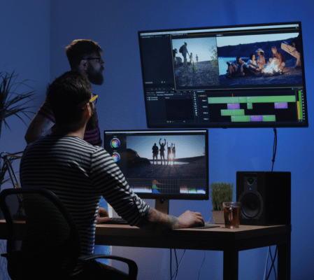 Two Video Editors Work In A Dimly Lit Room With Dual Monitors Displaying Video Editing Software. One Person, Seated At A Desk, Focuses On Color Correction While The Other Stands Nearby, Observing The Screens. A Potted Plant And A Speaker Sit On The Desk. Two video editors work in a dimly lit room with dual monitors displaying video editing software. One person, seated at a desk, focuses on color correction while the other stands nearby, observing the screens. A potted plant and a speaker sit on the desk.