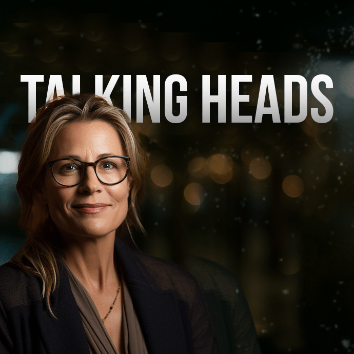 A person with shoulder-length hair and glasses is smiling slightly, dressed in a suit. The background is dark with blurred lights, and the title "TALKING HEADS" is prominently displayed above the person’s head.