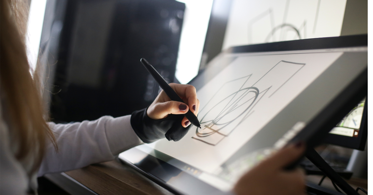 A person uses a digital pen to draw geometric shapes on a tablet screen, crafting intricate motion graphics for businesses. They wear gloves on one hand and are focused on creating the design. Nearby, computer equipment is visible, indicating a digital workspace.