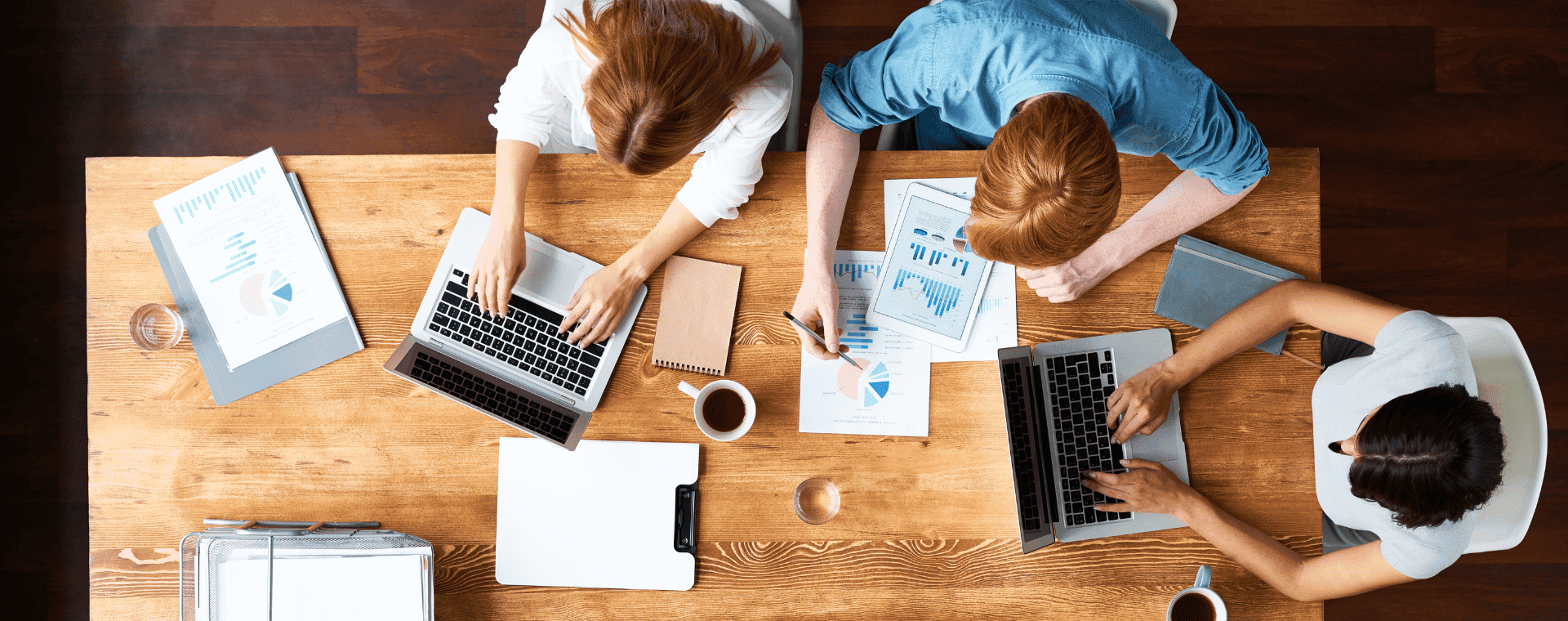 Three people are seated at a wooden table working on laptops. Papers with charts and graphs, two coffee cups, a notepad, a clipboard, and a binder are spread out on the table. The view from above showcases a collaborative office environment focused on b2b marketing strategies.