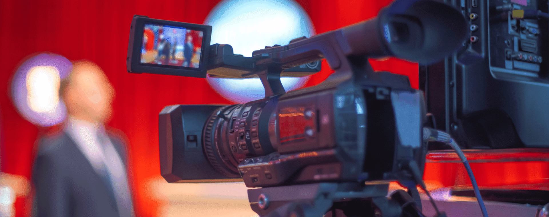 A close-up shot of a professional video camera pointed towards a speaker in a blurred background. The scene takes place in a brightly lit room with red curtains, and the camera's flip screen displays the speaker in focus—perfect for capturing engaging b2b marketing content.