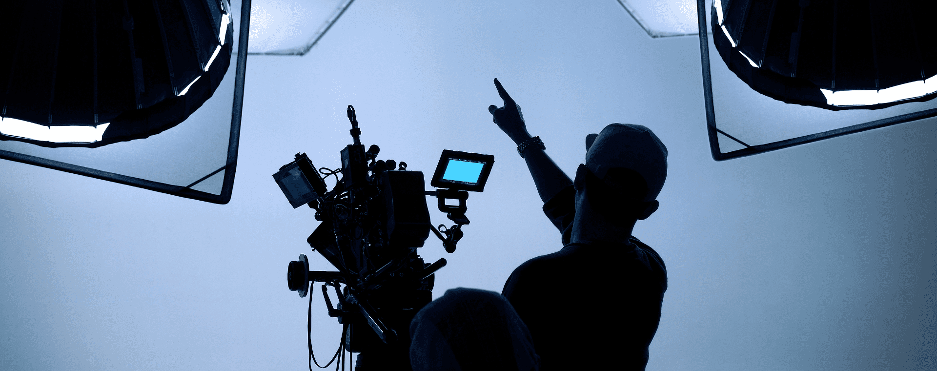 A silhouette of a person in a cap pointing upwards near a film camera setup with multiple monitors and lights in a studio environment. The backdrop, softly lit, creates a dramatic contrast with the equipment and the figure, evoking the precision often seen in b2b marketing visuals.