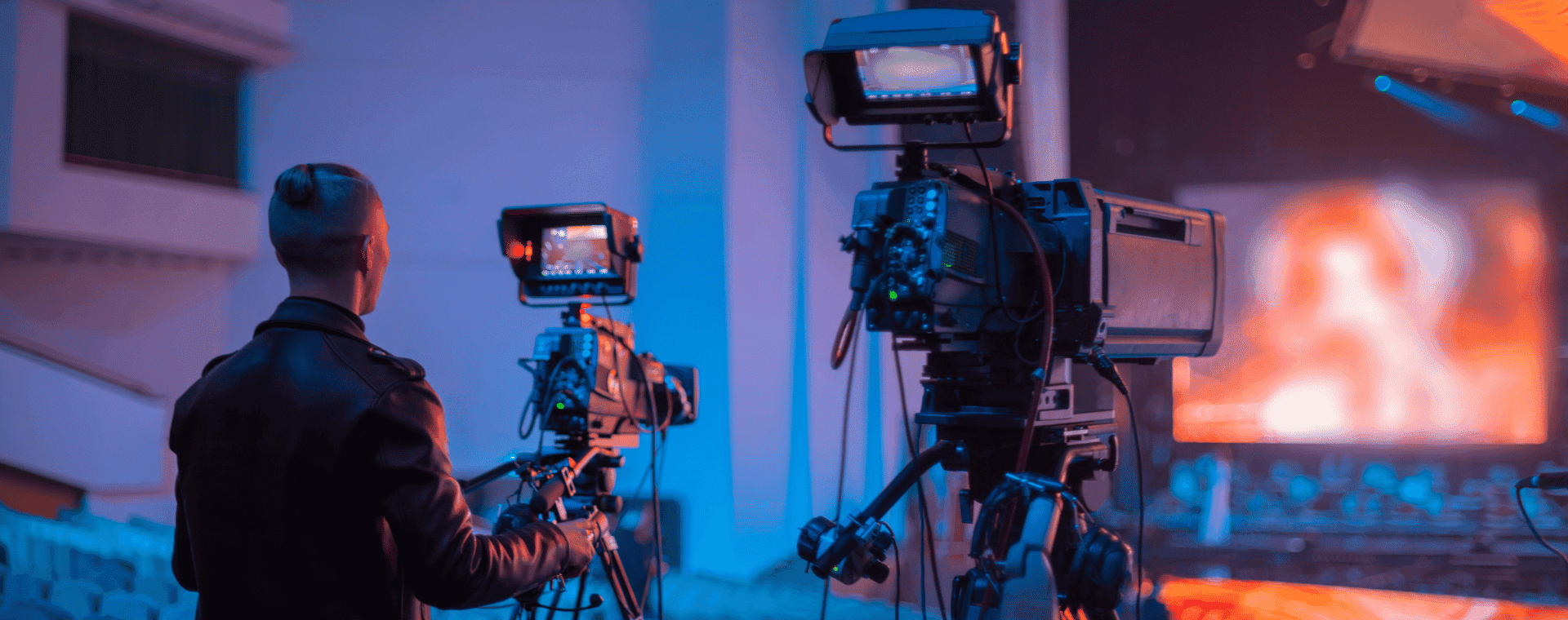 A person operates a professional video camera, capturing footage on a well-lit stage in a large auditorium. Multiple cameras are set up, with one prominently in the foreground. The scene, perfect for B2B marketing, is lit with blue and orange hues, and the seating area is visible in the background.